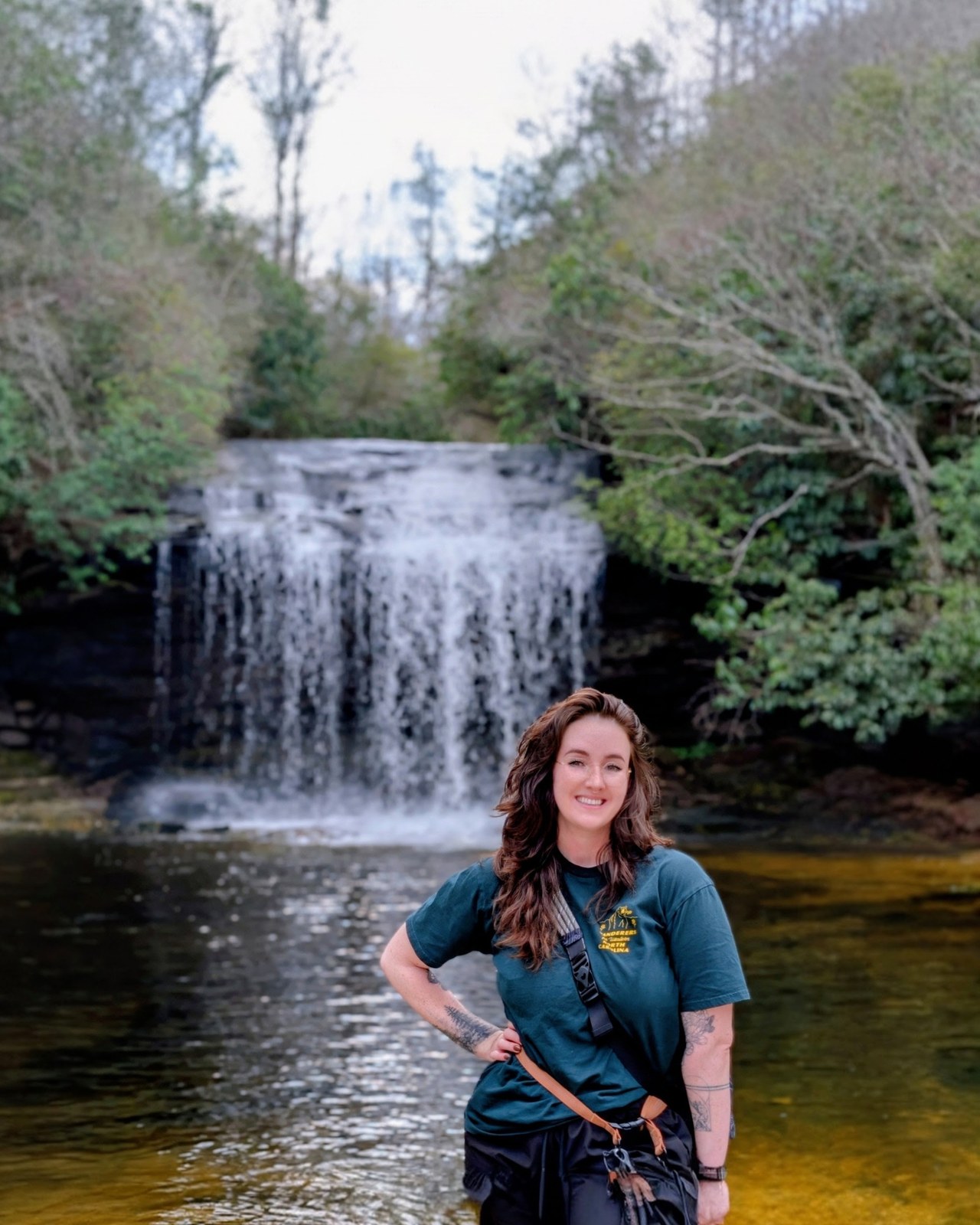Chantel Hill standing in a shallow river below a wide waterfall in a green Appalachian forest.